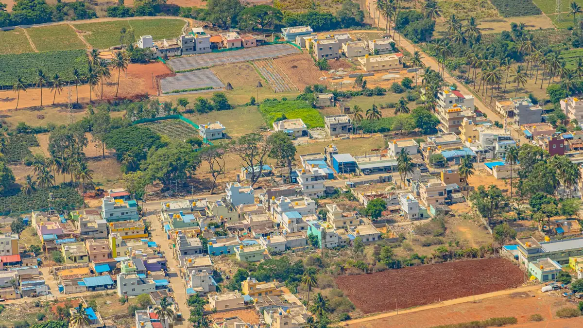 Aerial view of a growing residential layout at the edge of a town: rows of new two- and three-storey houses among coconut palms, with open red-earth plots and green farm fields around them
