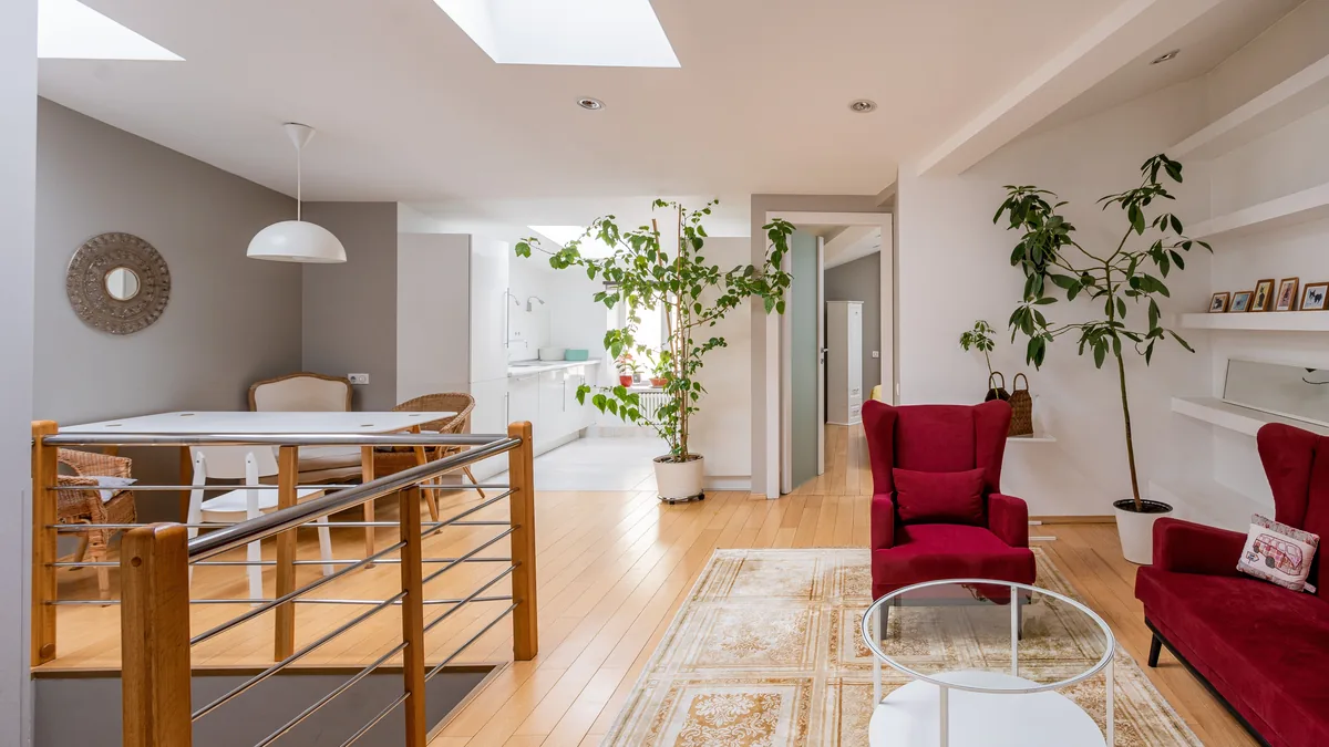 An open-plan living and dining space lit by skylights, with a pale wood floor, white dining table, deep red velvet armchairs and tall indoor plants